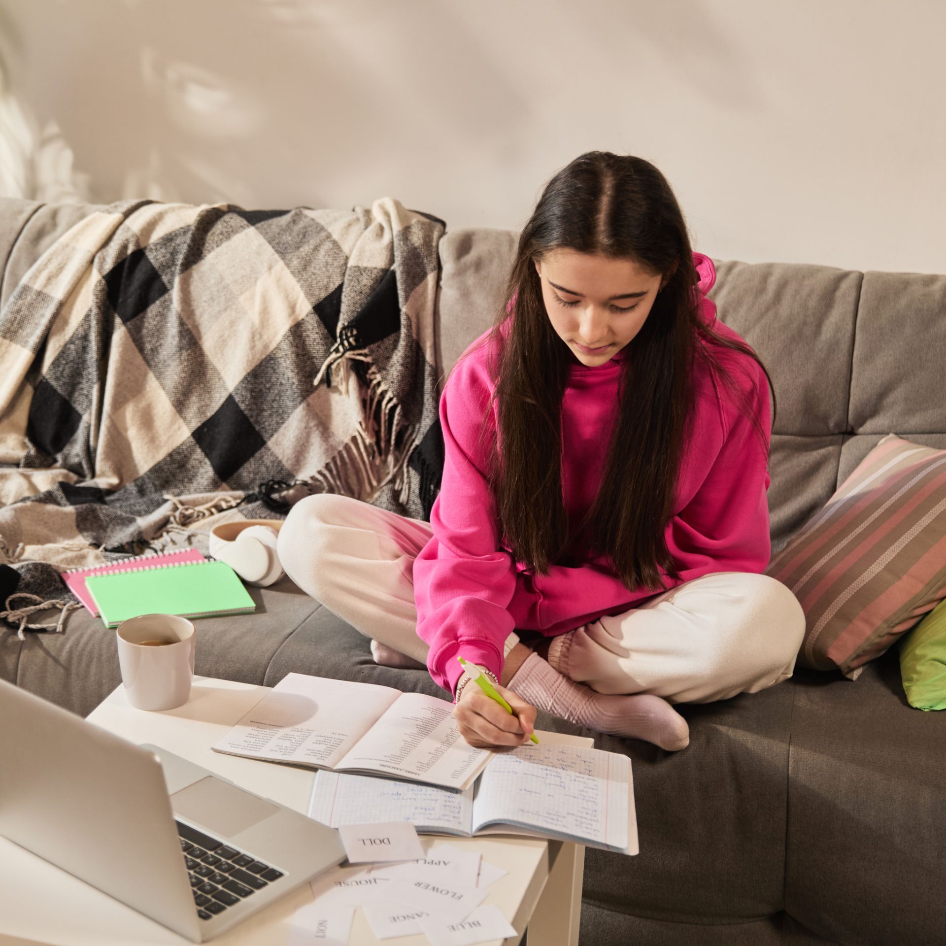 Girl in pink hoodie sitting on couch with open notebooks and laptop, writing English vocabulary in study journal. Teen girl studying 244184542 m scaled