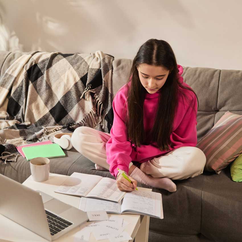 study-skills-exam-revision-coaching-girl-in-pink Girl in pink hoodie sitting on couch with open notebooks and laptop, writing English vocabulary in study journal. Concept of education, homeschooling, studying. Online lesson with tutor